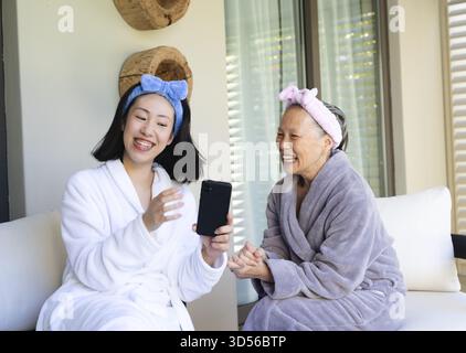 Asiatisch-chinesische Mutter und Tochter lachen und teilen ihr Smartphone auf einem Terrassensofa mit Spa-Kopfbügeln Stockfoto