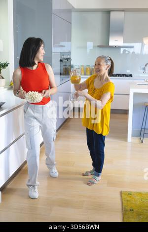 Asiatische Mutter und Tochter gießen Orangensaft, halten eine Schüssel Popcorn in der Küche, mit Glasgläsern Stockfoto