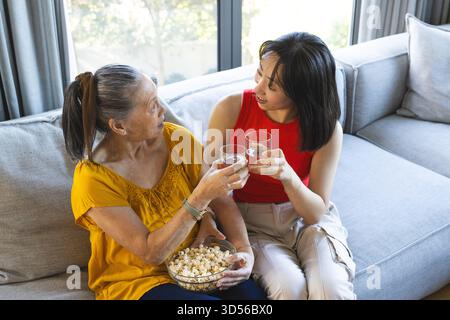Plaudernde asiatische Mutter und Tochter halten klare Gläser in hellem Wohnzimmer, mit Popcorn-Schüssel Stockfoto
