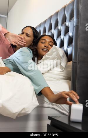 Zwei gähnende Frauen, die im Schlafzimmer liegen, mit weißer Bettwäsche und dunklem Kopfteil Stockfoto