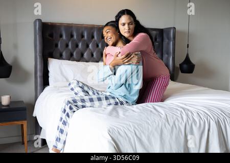 Sie umarmen verschiedene weibliche Freunde, die im modernen Schlafzimmer auf dem Bett sitzen, mit Keramikbecher und Pendelleuchten Stockfoto