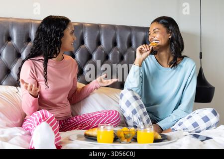 Sie unterhalten sich mit verschiedenen Freundinnen, die sich im Bett lachen, mit Frühstückstablett und Croissants Stockfoto
