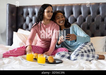 Lächelnde, vielfältige Freundinnen, die ihr Smartphone auf dem Bett halten, mit Saft und Croissants Stockfoto