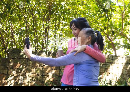 Lächelnde asiatische Großmutter und Enkelin machen Selfie im Garten, mit Smartphone und Getränk Stockfoto