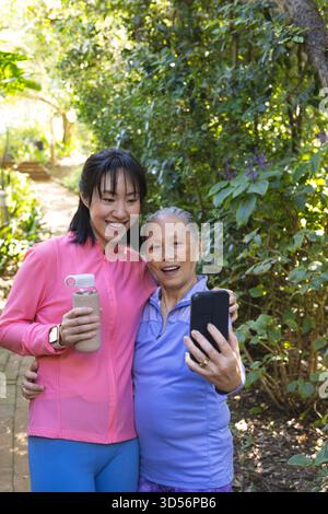 Lächelnde chinesische Mutter und Tochter machen Selfie auf dem Gartenweg, mit Smartphone und Wasserflasche Stockfoto