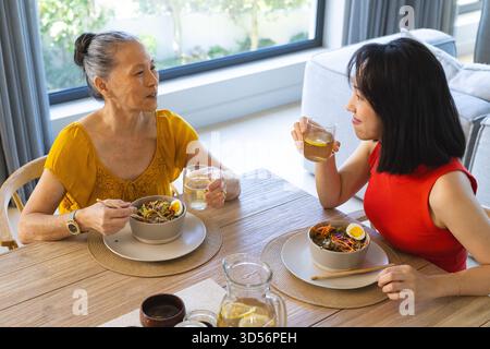 Asiatische Mutter und Tochter sitzen zu Hause am Esstisch, mit Nudelsalat und Getränken Stockfoto