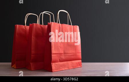 Packung mit drei leeren roten Papiertaschen mit braunen, verdrehten Papiergriffen auf einem Holztisch und dunklem, isoliertem Hintergrund. Vorderansicht. Stockfoto