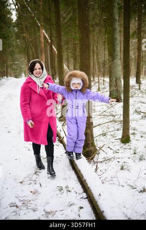 Die Mutter unterstützt das Kind, während es auf einem verschneiten Pfad auf umgestürzten Bäumen geht, sie beschützt, hält und ermutigt. Mutter hilft Mädchen, auf einem schneebedeckten Weg auf Moos zu balancieren. Winterspaziergänge im verschneiten Wald Stockfoto
