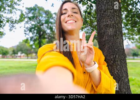 Lächelnde Frau in gelbem Hemd, Selfie im Freien, mit Friedenszeichen, Park. Stockfoto