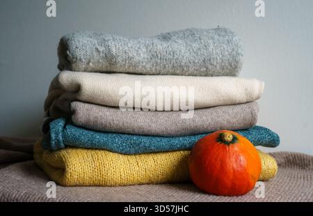 Ein Stapel weicher Strickpullover in neutralen Farben mit einem kleinen Kürbis sorgt für eine warme und gemütliche Herbstatmosphäre Stockfoto