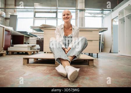 Glatze Frau lächelt im Overall, sitzt auf Palette in der Möbelwerkstatt Stockfoto
