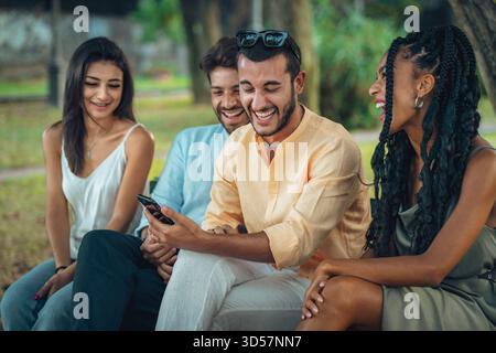 Eine fröhliche, multiethnische Gruppe von vier jungen Freunden sitzt auf einer Parkbank und lacht zusammen, während sie auf ein Smartphone schaut. Stockfoto