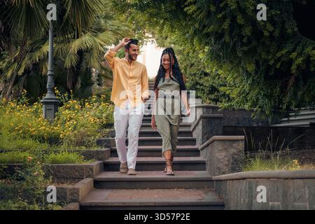 Ein stilvolles und glückliches Multi-ethnisches Paar geht eine Steintreppe in einem wunderschönen grünen Park hinunter und lächelt zusammen in einem Sommerurlaub. Stockfoto