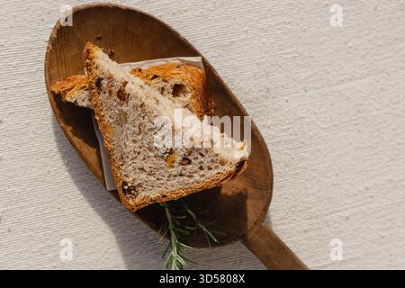 Zwei Scheiben handwerkliches getrocknetes Fruchtbrot auf rustikalem Holzbrett, roher Leinentischdecke, Rosmarin. Traditionelle Bäckerei, Brot, rustikal, authentisch Stockfoto