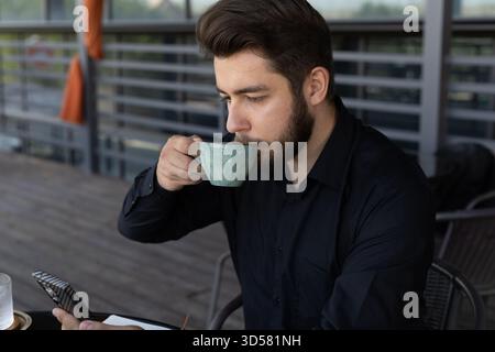 Ein bärtiger Mann sitzt und trinkt aus einer Tasse, während er auf sein Handy schaut. Stockfoto