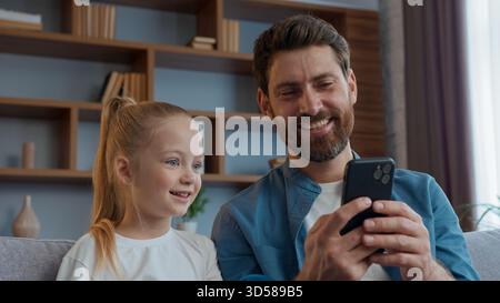Kaukasische glückliche Familie aussehendes Telefon lachender Vater mit Tochter lachen mobiles Video lustige Inhalte nettes Kind Mädchen Kind Blick auf Smartphone Stockfoto