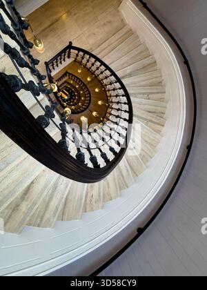 Die Wendeltreppe von oben mit kunstvollen schwarzen Geländern, warmen Pendelleuchten und Marmortreppen sorgt für eine elegante architektonische Atmosphäre Stockfoto