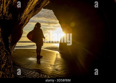 Blick auf eine Silhouette einer Person mit einem Surfbrett, das in einem Höhleneingang steht, mit Sonnenlicht, das vom Meer reflektiert wird, und nassem Sand, Tongaporutu, Tarana Stockfoto