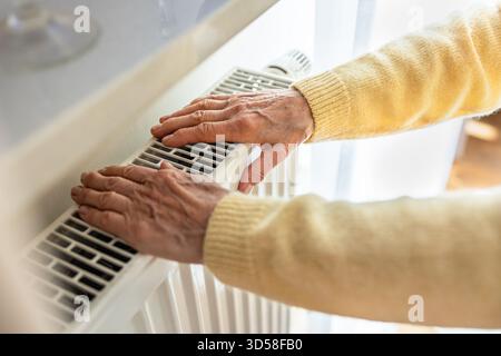 Eine ältere Frau wärmt ihre Hände auf einem Heizkörper zu Hause, Nahaufnahme Stockfoto