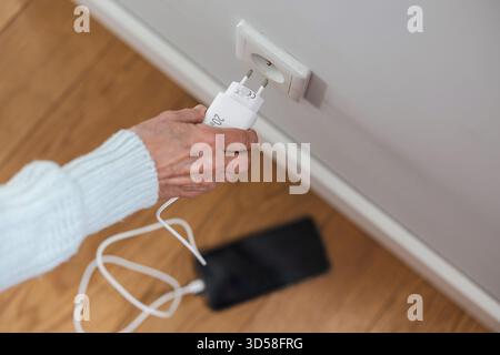 Nahaufnahme einer älteren Frau, die einen Netzstecker in eine Steckdose steckte, um ihr Telefon aufzuladen Stockfoto