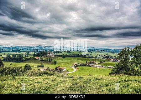 Panoramablick auf die Burgruine Sulzberg im Oberallgäu, Bayern. Die mittelalterlichen Überreste erheben sich über grünen Wiesen und ländlichen Bauernhöfen, umgeben von dramatischen Wolken und nebeligen Hügeln im Hintergrund. Stockfoto