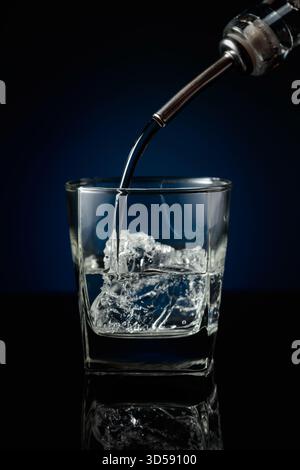 Kristallklarer Wodka oder Gin wird über Eis in ein Glas gegossen. Wodka mit Eis auf schwarzem reflektierendem Hintergrund. Stockfoto