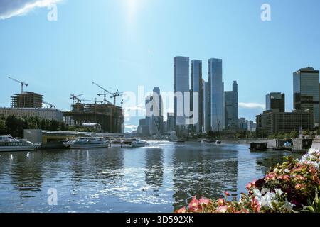 25. August 2025, Moskau, Russland. Wolkenkratzer erheben sich über einem ruhigen Fluss, der das Sonnenlicht reflektiert, während die Boote durch das Wasser fahren, umgeben von Konst Stockfoto
