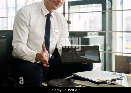 Kaukasier mittleren Alters in Geschäftskleidung, der am Schreibtisch des Autohauses sitzt und ein Tablet mit Innenbild des Autos präsentiert, Gesten mit Hand, Dokumente und Stift auf dem Tisch Stockfoto
