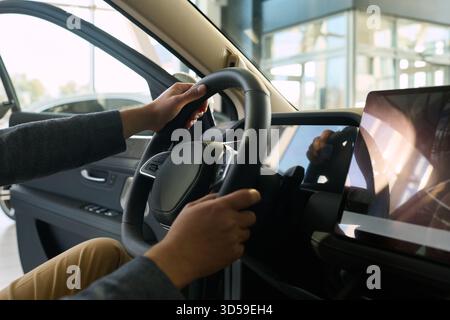Junger Erwachsener asiatischer Mann, der auf dem Fahrersitz sitzt und das Lenkrad hält, mit beiden Händen im modernen Auto im Händlerbetrieb, offene Tür und digitales Armaturenbrett im Vordergrund sichtbar Stockfoto