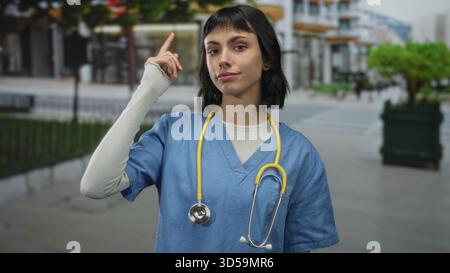 Krankenschwester mit blauem Peeling und Stethoskop zeigt den Finger auf den Tempel auf der Straße; Neugier reflektiert. Stockfoto
