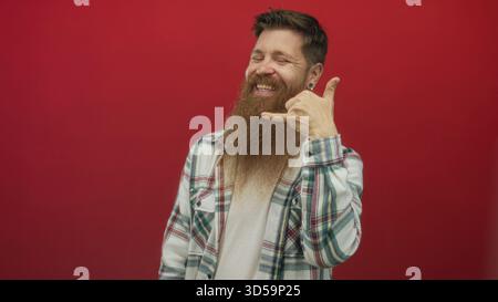 Junger bärtiger Mann mit kariertem Hemd und Ohrring, der im roten Studio Telefongeste macht; Kommunikationswärme. Stockfoto
