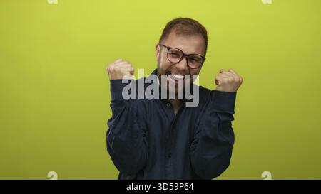Mann mit geballten Fäusten und lachender Brille, in einem hellgrünen Atelier in einem marineblauen Hemd; Glücksfeier Erfolg. Stockfoto