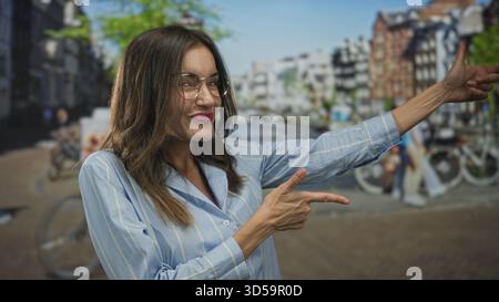 Lächelnde Frau in blauem Hemd mit Brille und breitem Grinsenfinger, der auf den sonnendurchfluteten Kanal auf der Straße zeigt; unbeschwerte Freude. Stockfoto