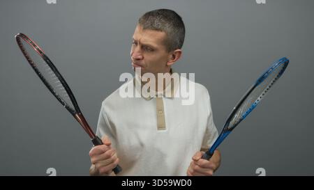 Mann in weißem Hemd, der zwei Tennisschläger vor isoliertem grauen Hintergrund hält, bewertet Ausrüstung mit einem durchdachten Ausdruck, der auf Deci hinweist Stockfoto