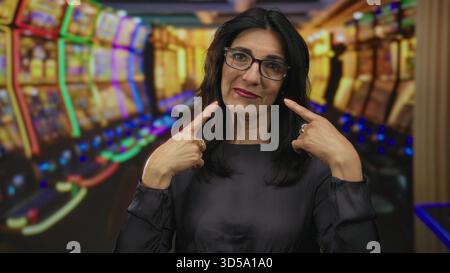 hispanische Frau mittleren Alters zeigt Finger auf Wangen neben leuchtenden Spielautomaten-Reihen im Casino; Freude. Stockfoto