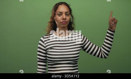 Die Frau zeigt den Zeigefinger im Studio nach oben und trägt ein gestreiftes Hemd vor grünem Hintergrund. Stockfoto