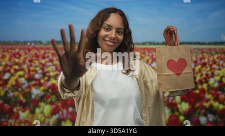 Junge Frau, die Hand winkt und einen Papiergeschenksack mit rotem Herzen in einem Tulpenfeld unter blauem Himmel hält; Freude Liebe Großzügigkeit. Stockfoto