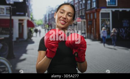 Die Frau hält Boxhandschuhe in einer Straßenboxhaltung hoch, lächelt und posiert mit roten Fäusten; Selbstbewusstsein stärkt. Stockfoto