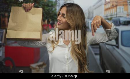 Junge blonde Frau hält Papiertüte und zeigt Finger an Tasche auf einer belebten Stadtstraße; Marketing Promotion Verkauf Shopping. Stockfoto