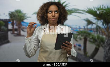 Junge afroamerikanische Bäckerin in Schürze, die Tablette hält und Finger auf der Straßenpromenade am Meer mit Palmen drückt; Präzision Stockfoto