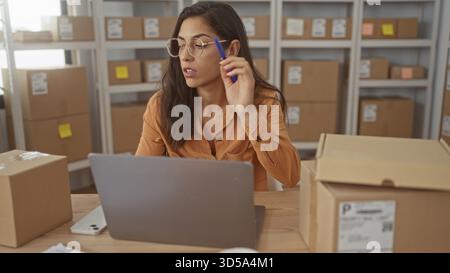Frau, die Pakete an einem Laptop überprüft, während sie einen Stift hält und von Kartonschachteln in einem Gebäude umgeben ist; Konzentration kleiner Unternehmen. Stockfoto