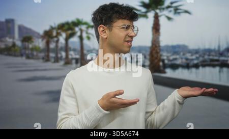 Mann mit weißem Pullover und Brille zeigt Palmen, die neben dem Boot auf der Straßenpromenade unter Palmen zeigen; Unsicherheit. Stockfoto