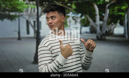 Ein junger Mann im Freien, der auf einer Stadtstraße mit Bäumen und Gebäuden im Hintergrund die Daumen hochlegt, ein gestreiftes Hemd trägt und selbstbewusst lächelt, verschwindet Stockfoto