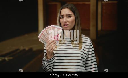 Frau mit isländischen Kronen-Banknoten auf einer Innenbaustelle mit einem verwirrten Ausdruck, der den unvollendeten Hintergrund kontrastiert. Stockfoto