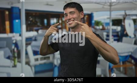 Der Mann zeigt Finger an Wange auf einer sonnendurchfluteten Restaurantterrasse auf einem Dach des Gebäudes mit Blick auf die Stadt; Glück. Stockfoto