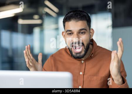Junger indischer Geschäftsmann, der überrascht und glücklich aussieht, während er etwas tolles auf einem Laptop sieht, während er sich in einem modernen Büro aufgeregt die Hände hebt Stockfoto