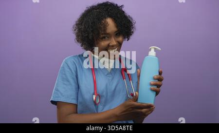 Frau Ärztin in blau schrubbt lächelnd hält Plastikpumpe Flasche mit Händen gegen violette Studiowand; Pflege. Stockfoto