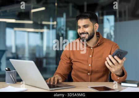Lächelnder junger Mann mit Bart, der an einem Holzschreibtisch in einem modernen Büro sitzt, Multitasking durch Tippen auf einem Laptop und Halten eines Smartphones, was Konnektivität und Produktivität repräsentiert Stockfoto