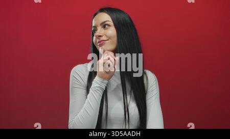 Junge hispanische Frau in weißem Hemd, die vor leuchtend roten Wänden posiert isolierter Hintergrund mit durchdachtem Ausdruck, der modernen Stil und Persönlichkeit zeigt Stockfoto