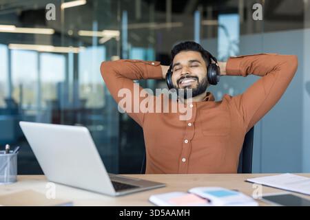 Junger Mann, der Kopfhörer trägt und sich mit den Händen hinter dem Kopf zurücklehnt, Musik genießt und Zufriedenheit findet, während er in einer modernen Büroumgebung eine Pause von der Arbeit macht Stockfoto